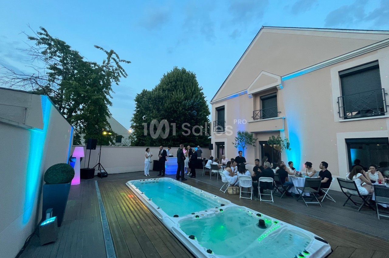 Personnes réunies autour de tables sur une terrasse avec un jacuzzi, devant une maison éclairée en soirée.