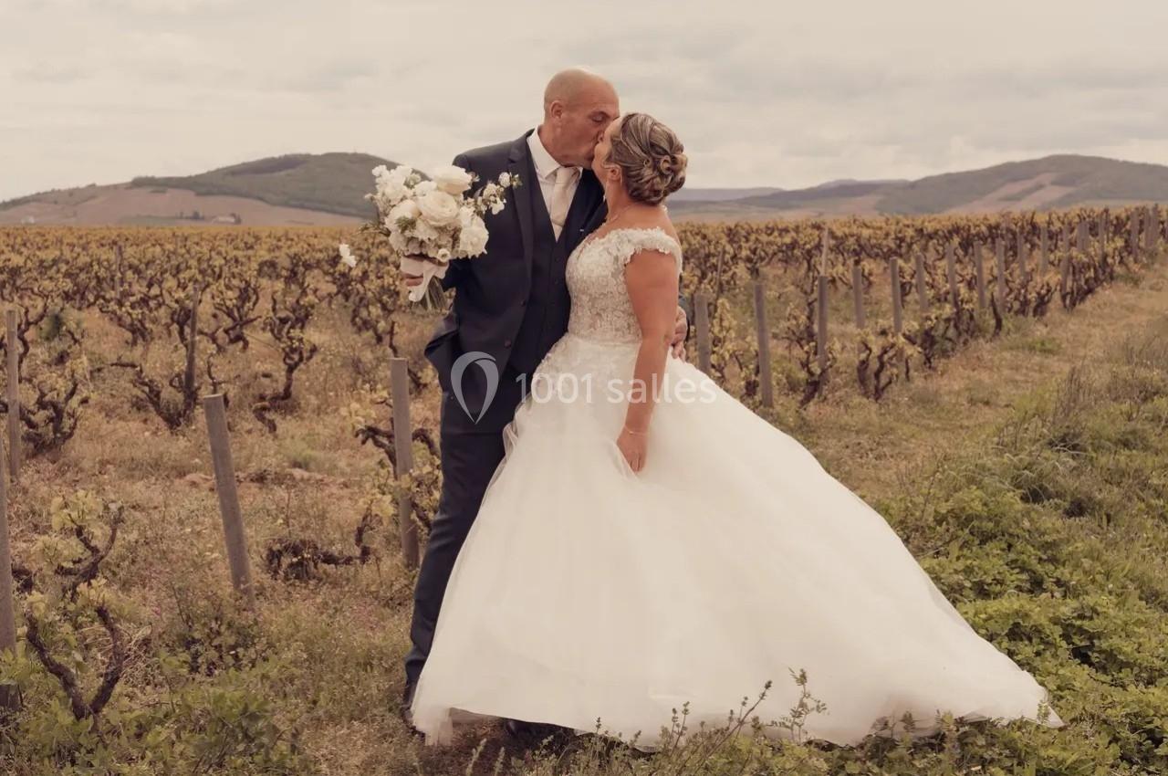 Un couple en tenue de mariage s'embrasse dans un vignoble sous un ciel nuageux.