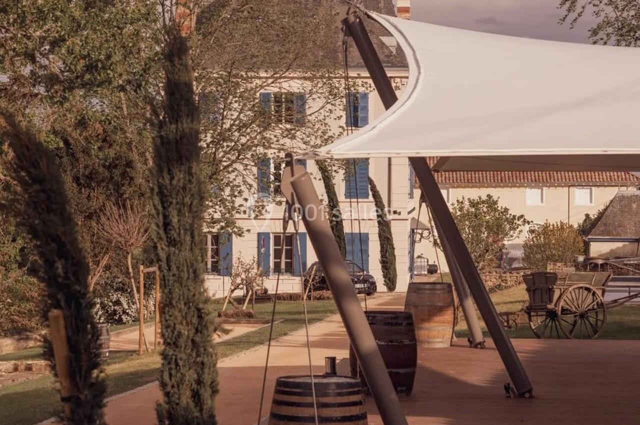Terrasse en bois avec des barriques, un voile d'ombrage et une maison en arrière-plan dans un cadre verdoyant.