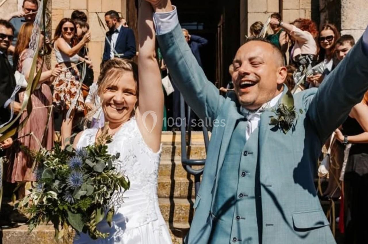 Un couple souriant en tenue de mariage sort d'un bâtiment, entouré d'invités célébrant avec joie.