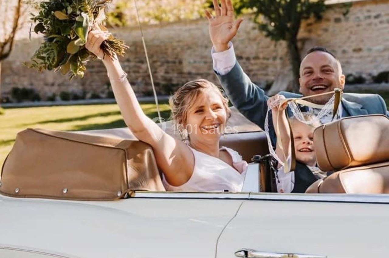 Un couple souriant avec un enfant dans une voiture décapotable, tenant des fleurs et saluant joyeusement.
