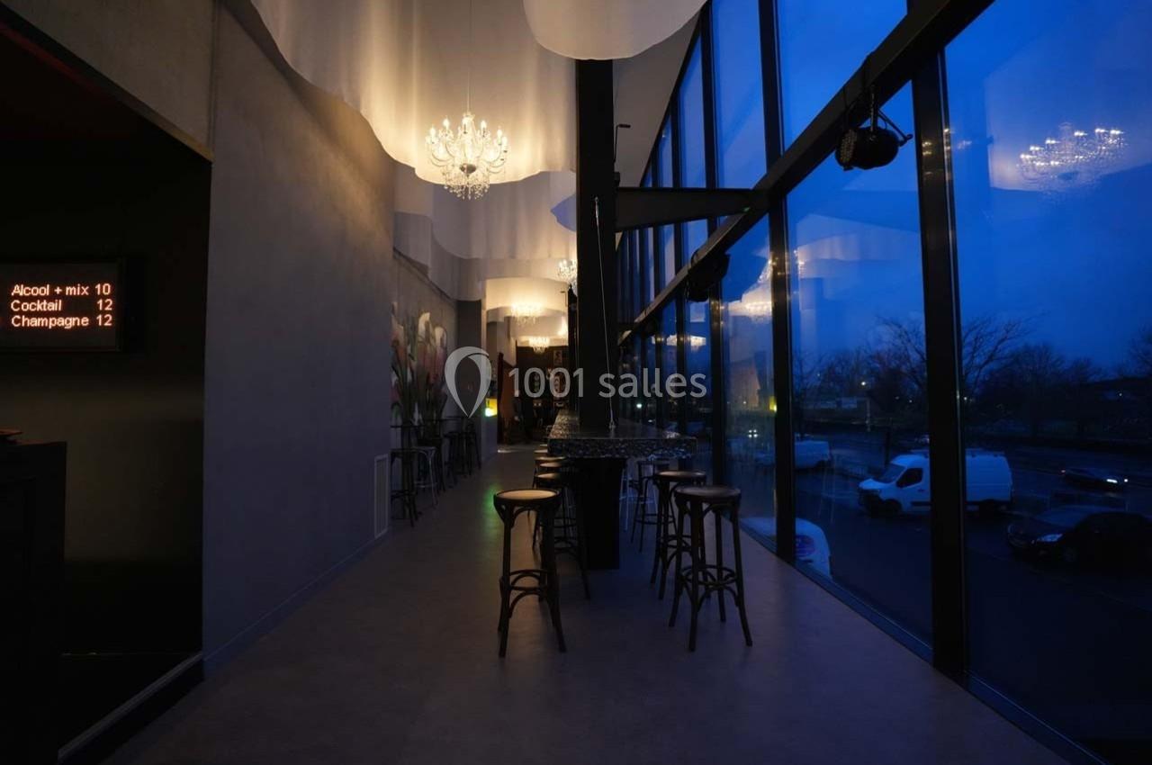 Intérieur d'un bar moderne avec tabourets hauts, grandes baies vitrées et éclairage tamisé en soirée.