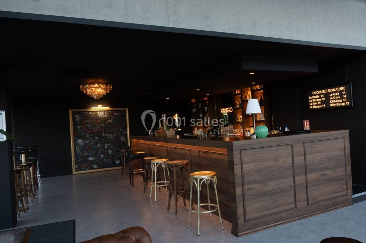 Bar moderne avec comptoir en bois, tabourets hauts, éclairage tamisé et étagères de bouteilles en arrière-plan.