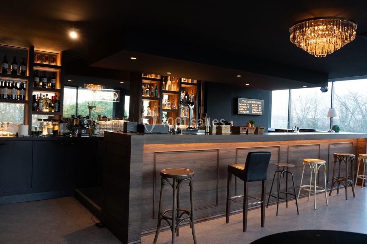 Bar moderne avec comptoir en bois, tabourets variés, étagères de bouteilles éclairées et lustre suspendu.
