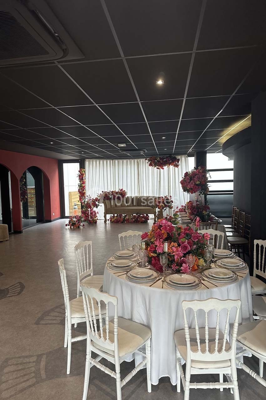 Salle de réception décorée avec des fleurs colorées, tables dressées avec nappes blanches et chaises assorties.