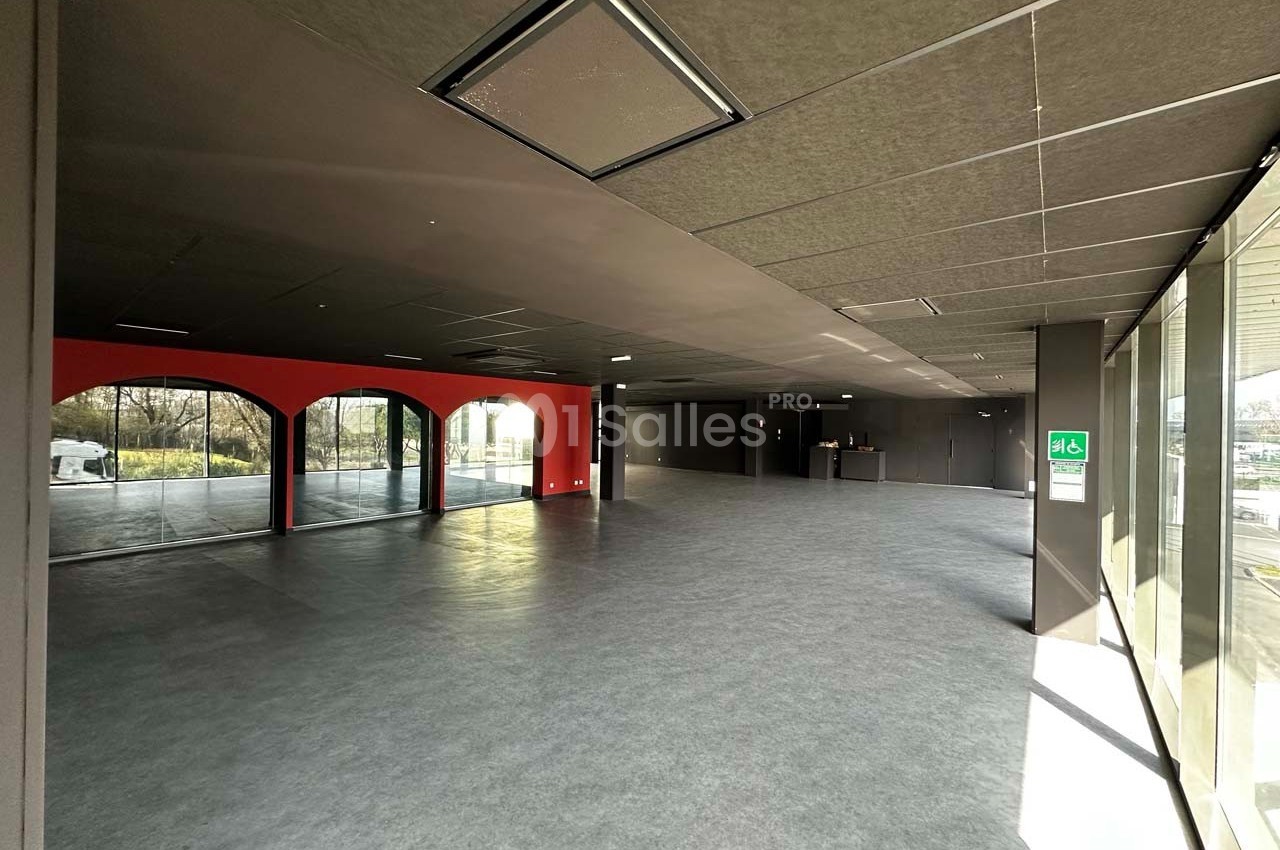 Intérieur d'un espace vide avec sol gris, murs sombres, grandes baies vitrées et lumière naturelle entrant.