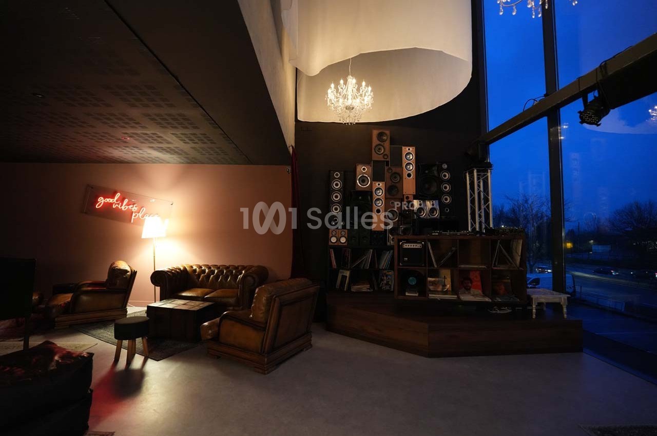 Salon cosy avec fauteuils en cuir, lampe, étagère de haut-parleurs et grande baie vitrée donnant sur l'extérieur.