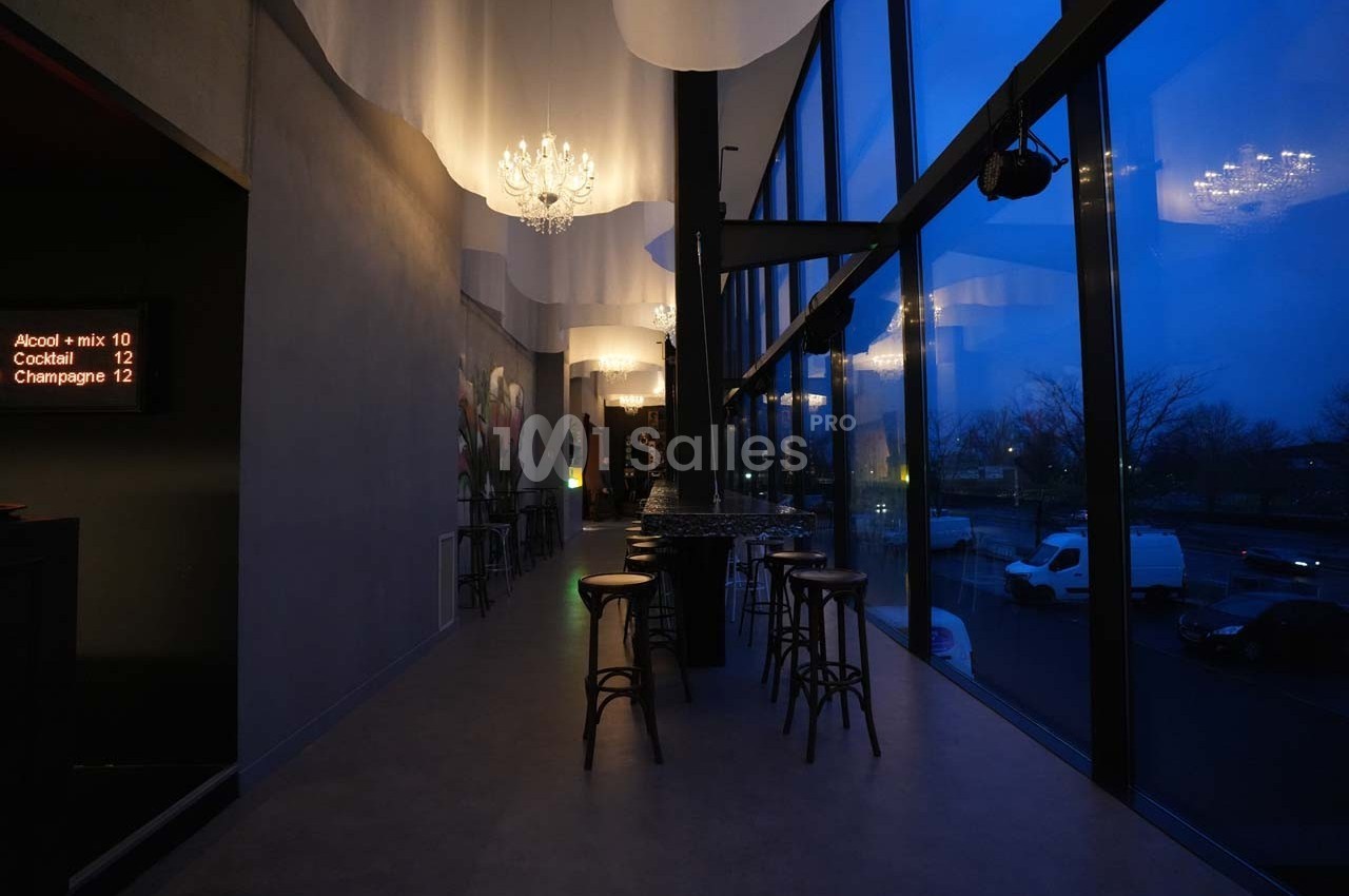 Intérieur d'un bar moderne avec tabourets hauts, grandes baies vitrées et éclairage tamisé en soirée.