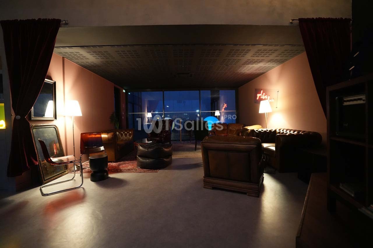 Salon cosy avec fauteuils en cuir, tapis rouge, lampes allumées et vue nocturne sur une grande baie vitrée.