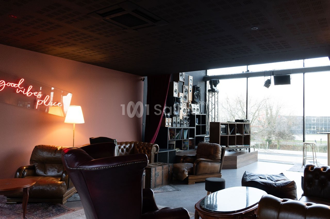 Salon moderne avec fauteuils en cuir, lampadaire, enseigne néon ’good vibes only’ et grande baie vitrée donnant sur l…