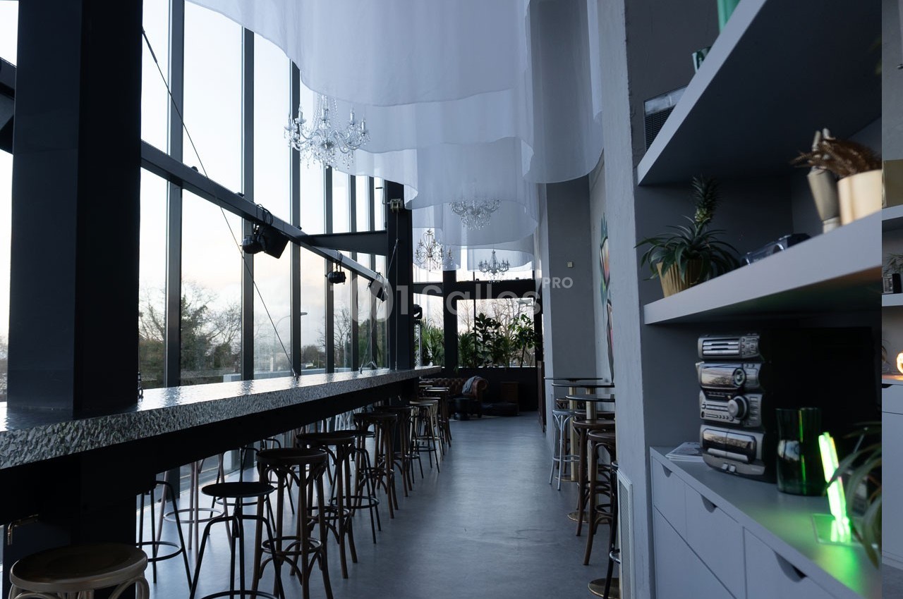 Intérieur d'un café moderne avec de grandes baies vitrées, un long comptoir, des tabourets et des étagères décorées.