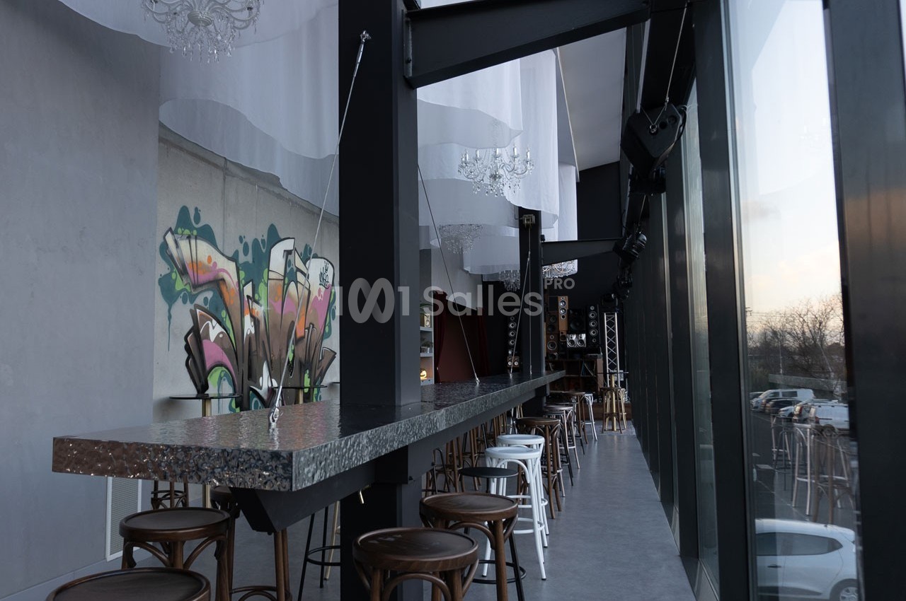 Intérieur d'un bar moderne avec un long comptoir métallique, des tabourets et une fresque murale colorée.