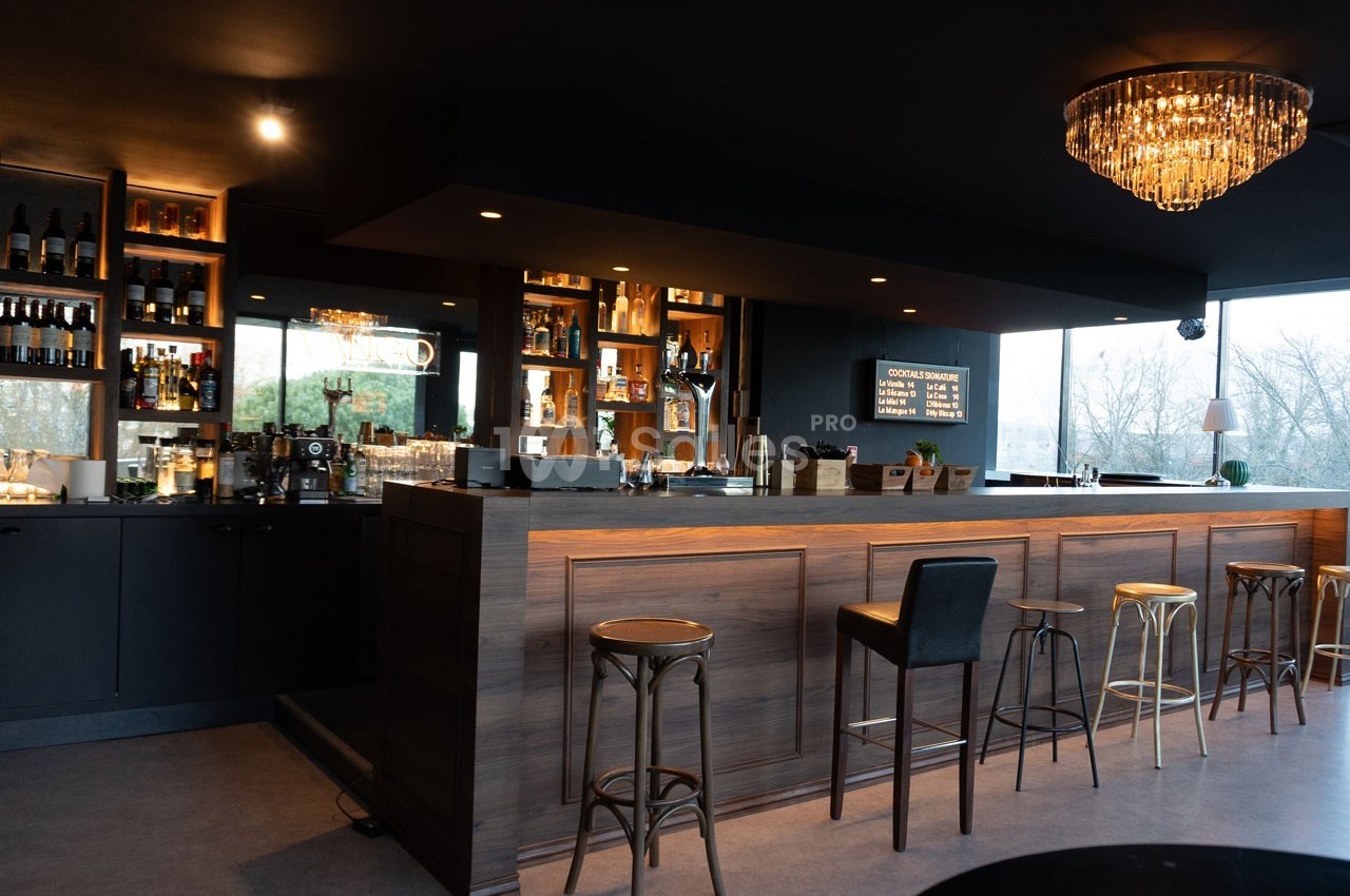Bar moderne avec comptoir en bois, tabourets variés, étagères de bouteilles éclairées et lustre suspendu.