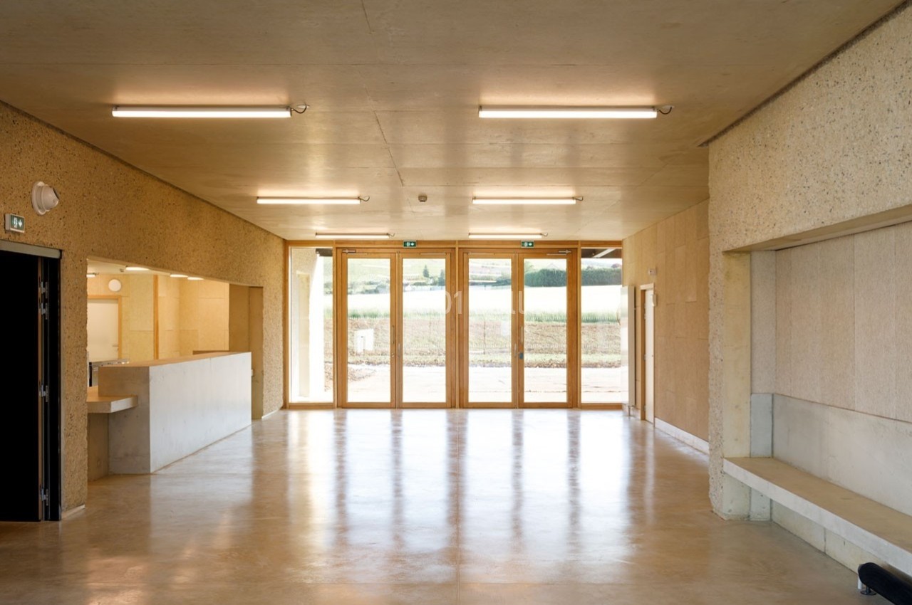 Salle lumineuse avec sol en béton, murs clairs, grandes baies vitrées et vue sur un paysage extérieur.