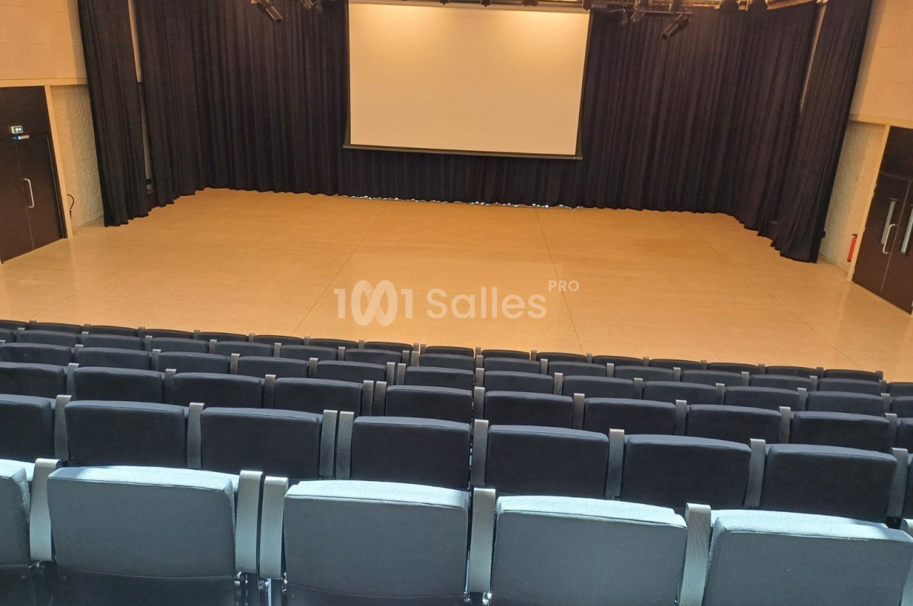 Salle de spectacle vide avec rangées de sièges noirs, scène en fond et écran blanc suspendu devant un rideau noir.