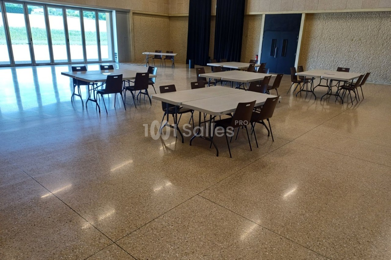 Salle avec sol en terrazzo, tables rectangulaires espacées et chaises, grandes baies vitrées et rideaux noirs en fond.
