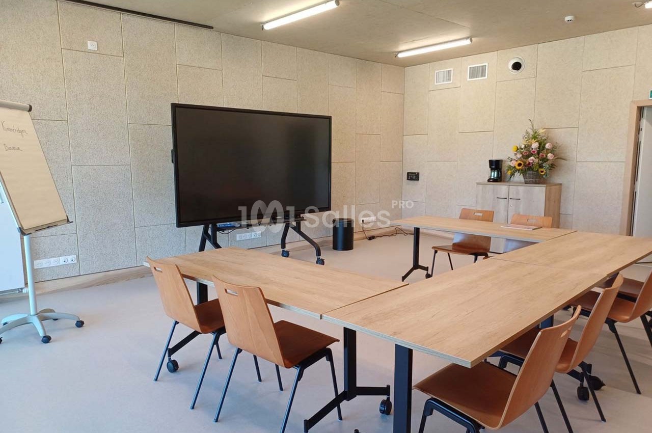 Salle de réunion lumineuse avec tables en bois, chaises, écran interactif, tableau papier et bouquet de fleurs.
