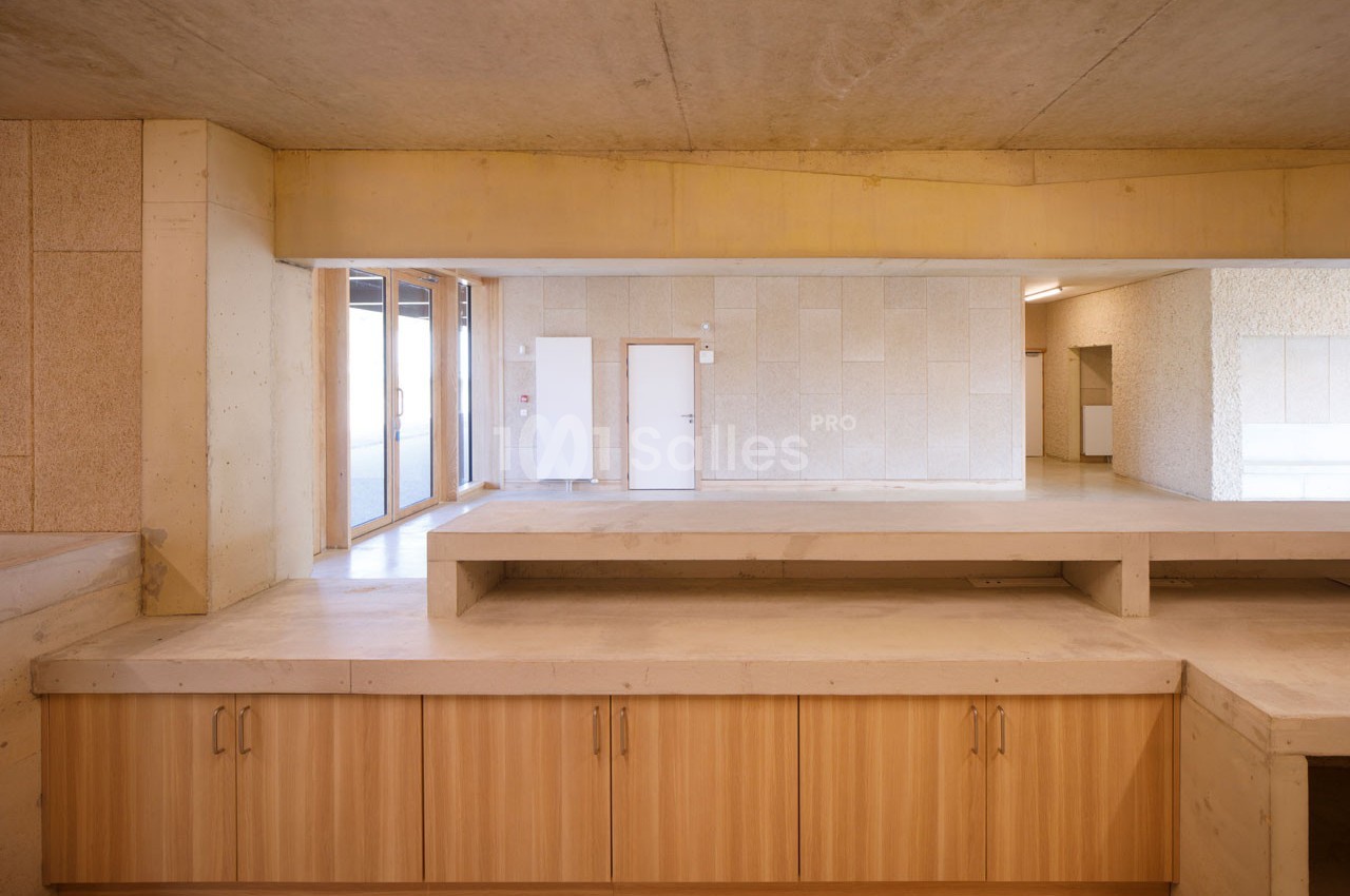 Intérieur d'un bâtiment avec murs en béton, mobilier en bois clair et lumière naturelle entrant par une baie vitrée.