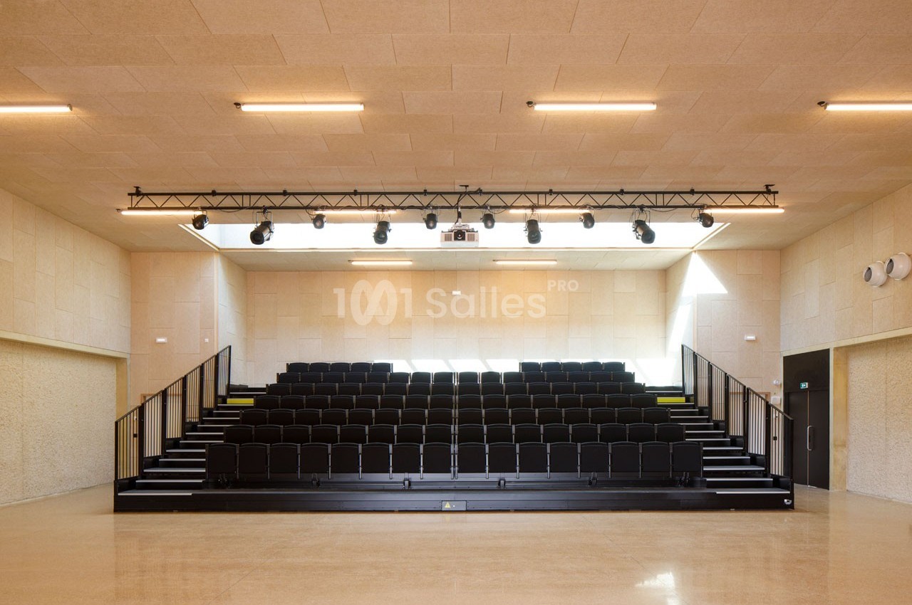 Gradins noirs vides dans une salle lumineuse avec un sol beige, des murs clairs et un éclairage suspendu.