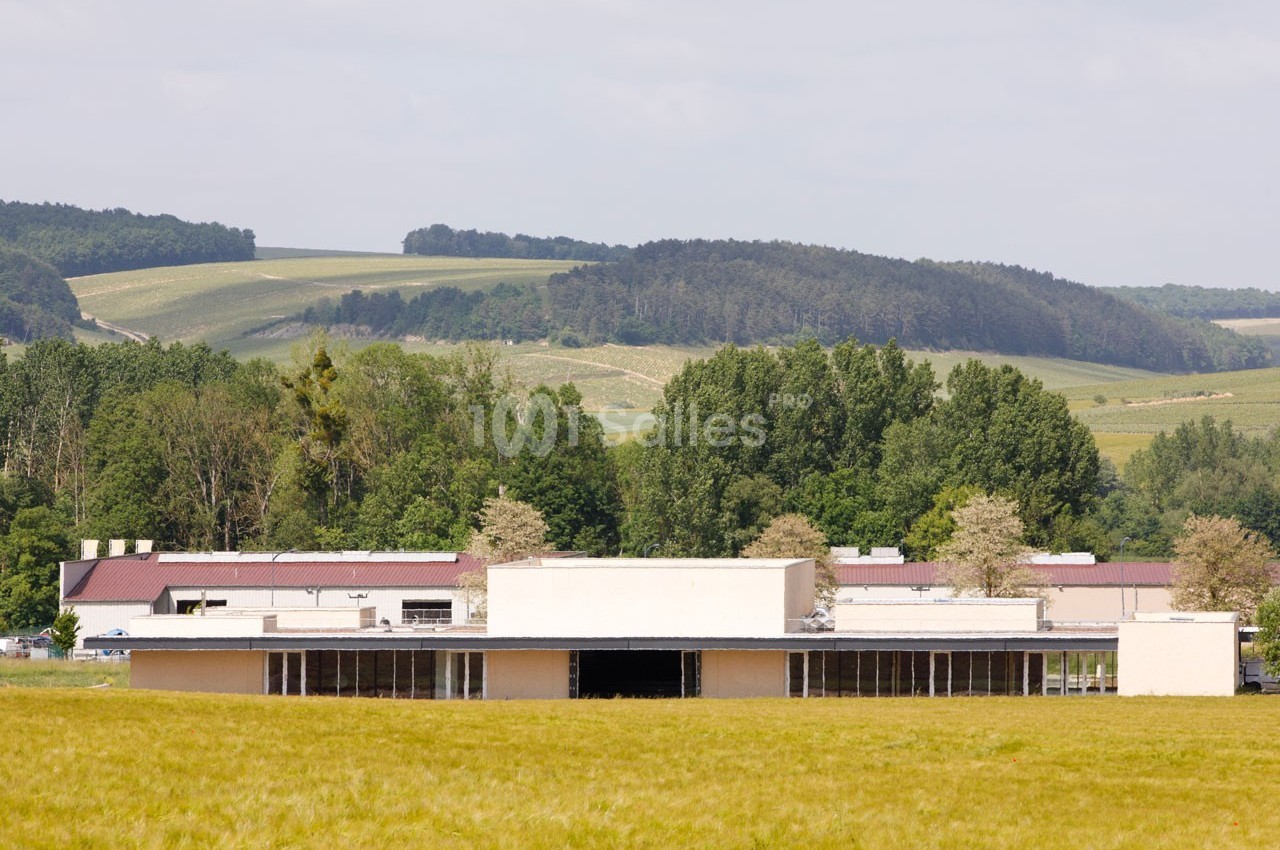 Bâtiment moderne entouré de champs et d'arbres, avec des collines boisées en arrière-plan sous un ciel clair.