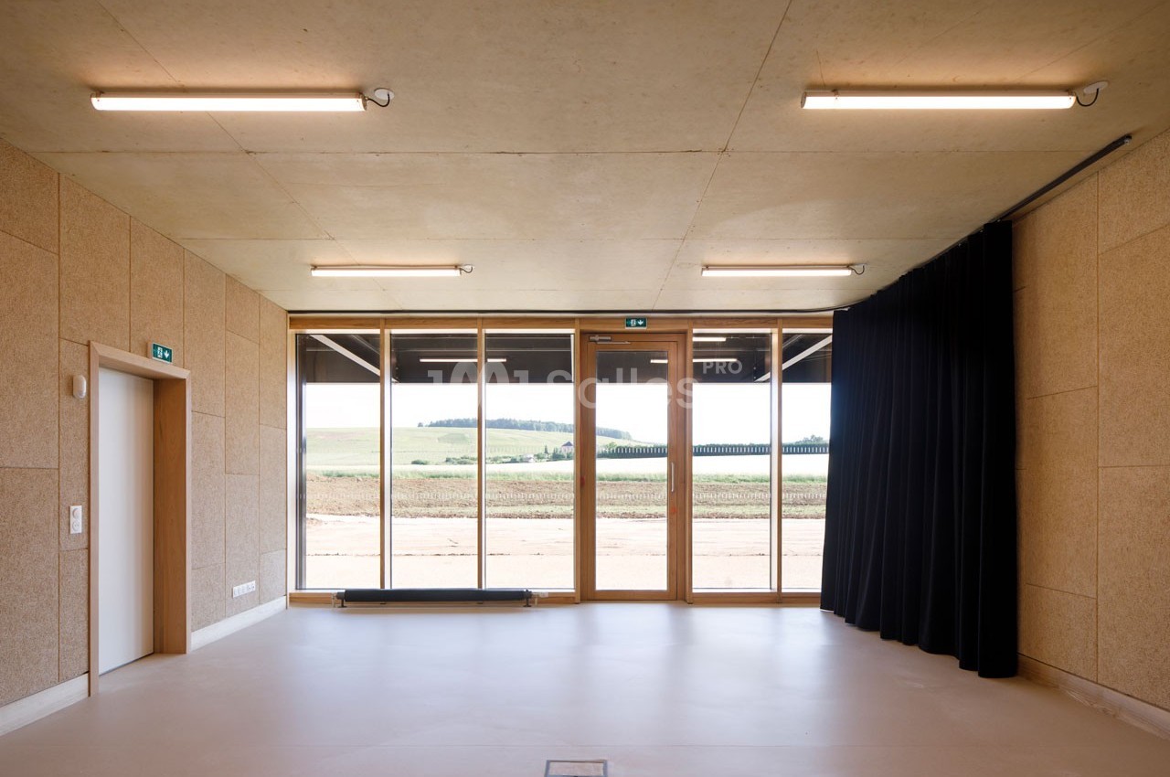 Salle lumineuse avec murs en bois, grandes baies vitrées donnant sur un paysage rural et rideaux noirs sur le côté.