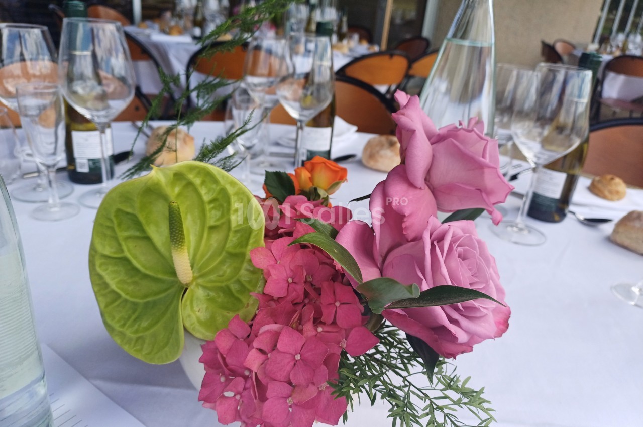 Centre de table floral avec roses roses, hortensias, anthurium vert et feuillage, entouré de verres et bouteilles.