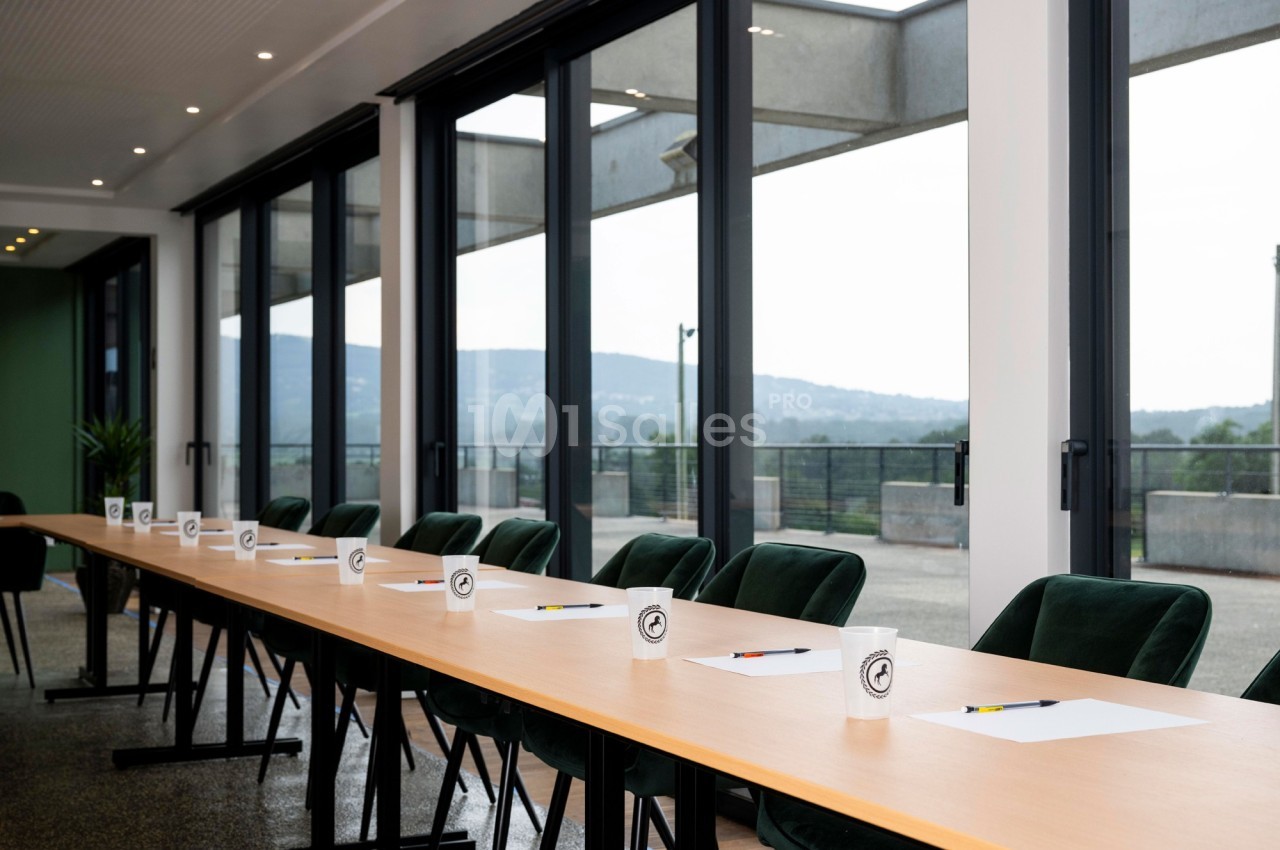 Salle de réunion lumineuse avec une longue table, des chaises vertes, des blocs-notes, des stylos et des gobelets alignés.