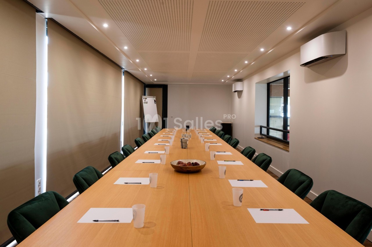 Salle de réunion avec une grande table en bois, chaises vertes, blocs-notes, stylos et carafe d'eau au centre.