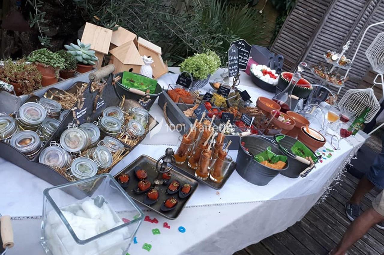 Buffet varié en extérieur avec verrines, plats colorés, boissons et décorations végétales sur une table blanche.