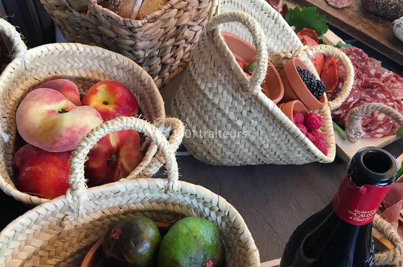 Paniers en osier contenant des fruits frais, avec charcuterie et bouteille de vin rouge en arrière-plan.