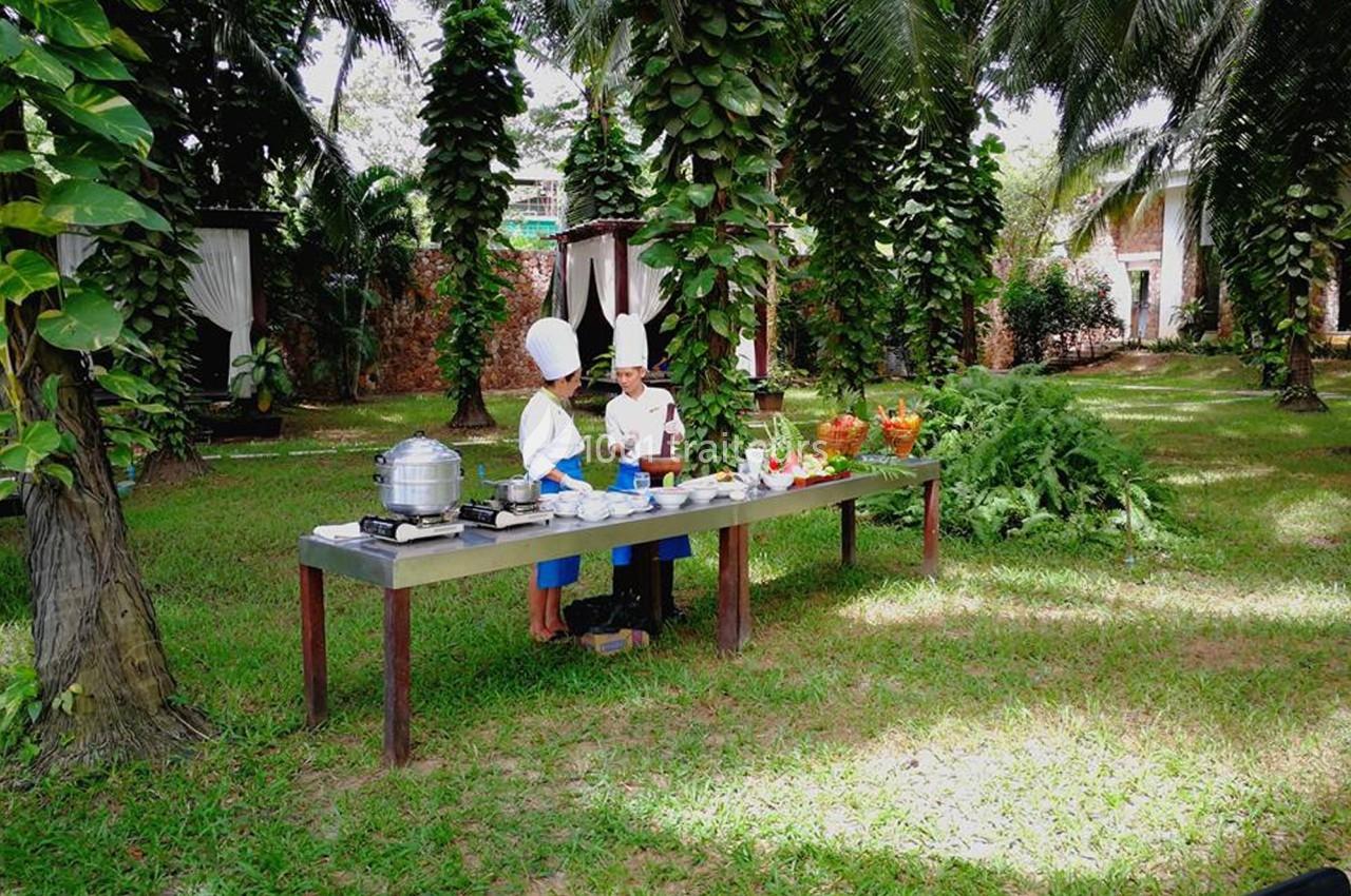 Deux chefs en tenue blanche préparent des plats sur une table en plein air, entourés de palmiers et de verdure.