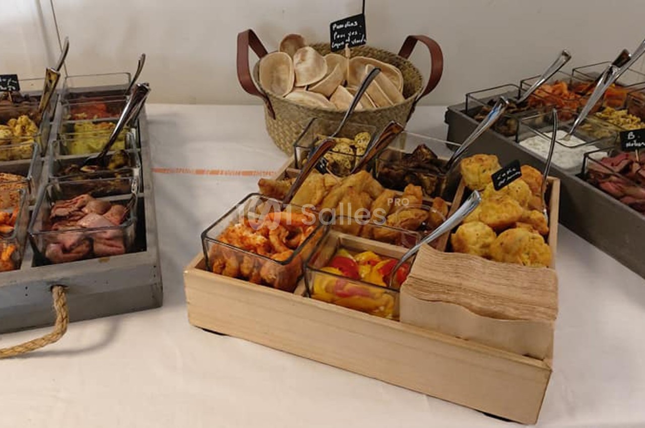 Buffet avec divers plats froids en verrines, pain pita dans un panier et serviettes en papier sur une table blanche.