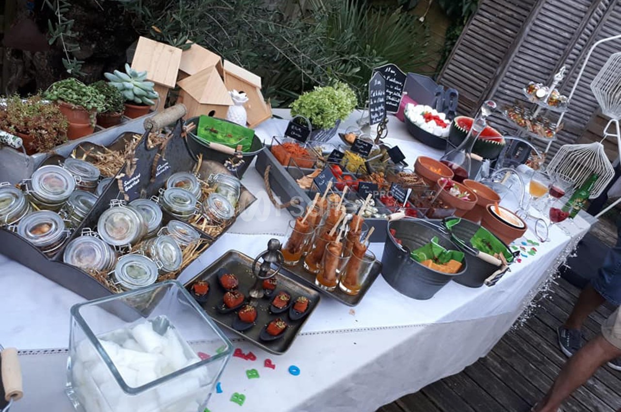 Buffet varié en extérieur avec verrines, plats colorés, boissons et décorations végétales sur une table blanche.