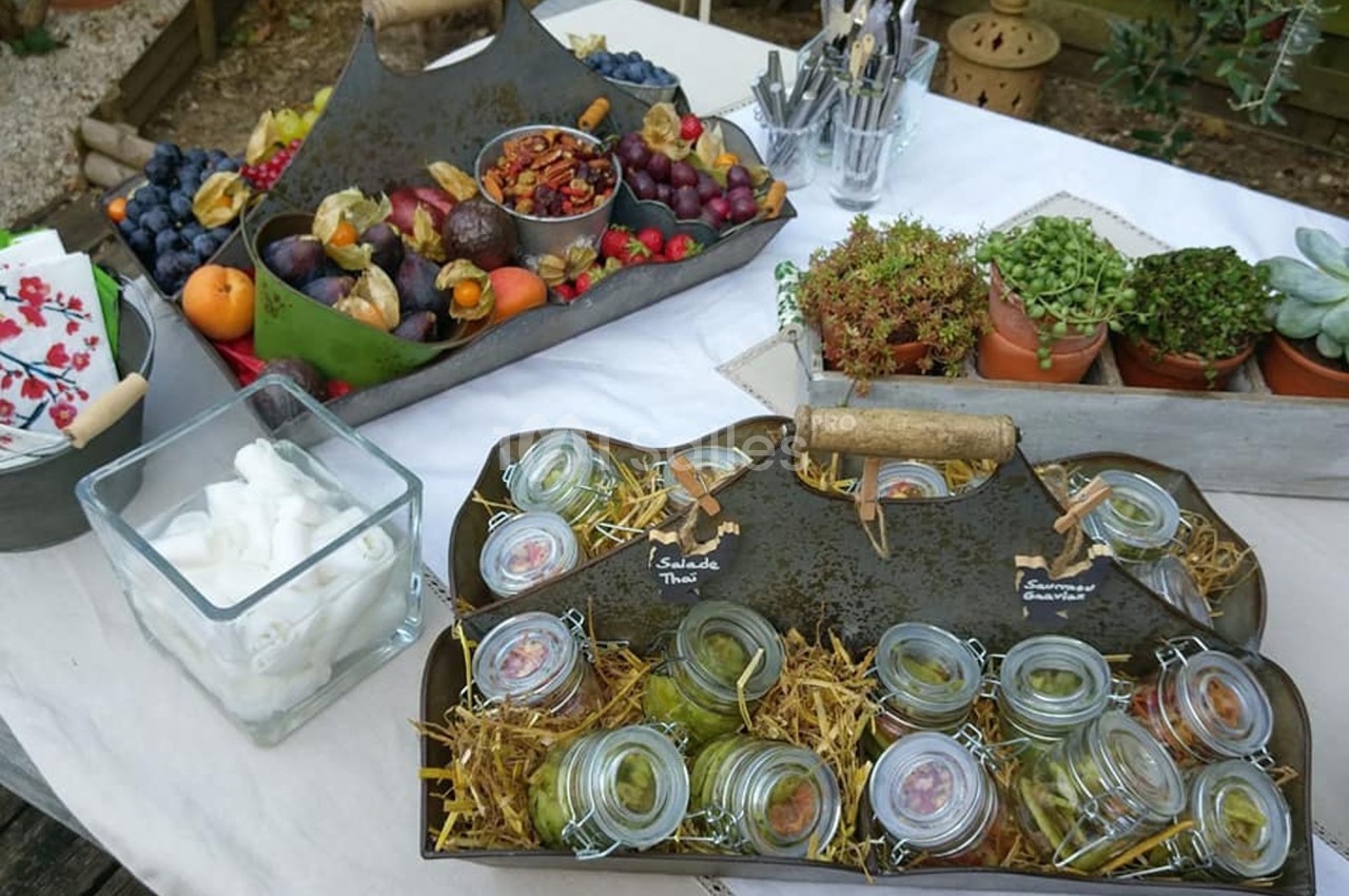 Plateau de bocaux en verre contenant des plats, entouré de fruits, serviettes et plantes sur une table extérieure.