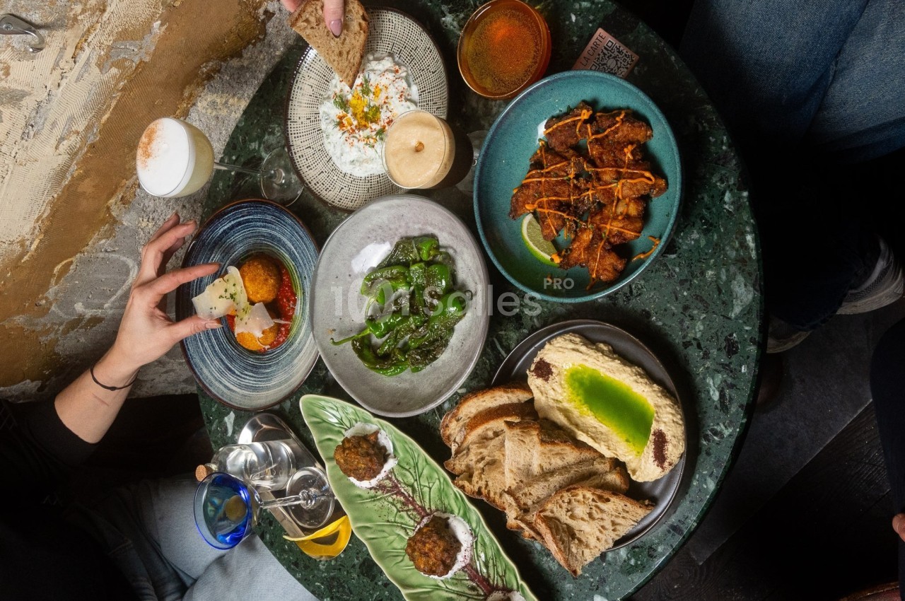 Table garnie de plats variés : pain, légumes, boulettes, sauces et boissons, avec des mains en train de servir.
