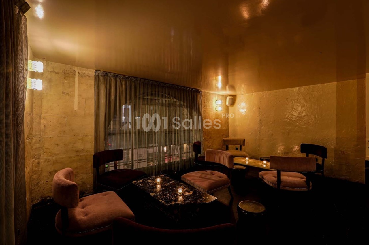 Salle intime avec éclairage tamisé, fauteuils en velours rose, table basse et rideaux donnant sur une fenêtre.