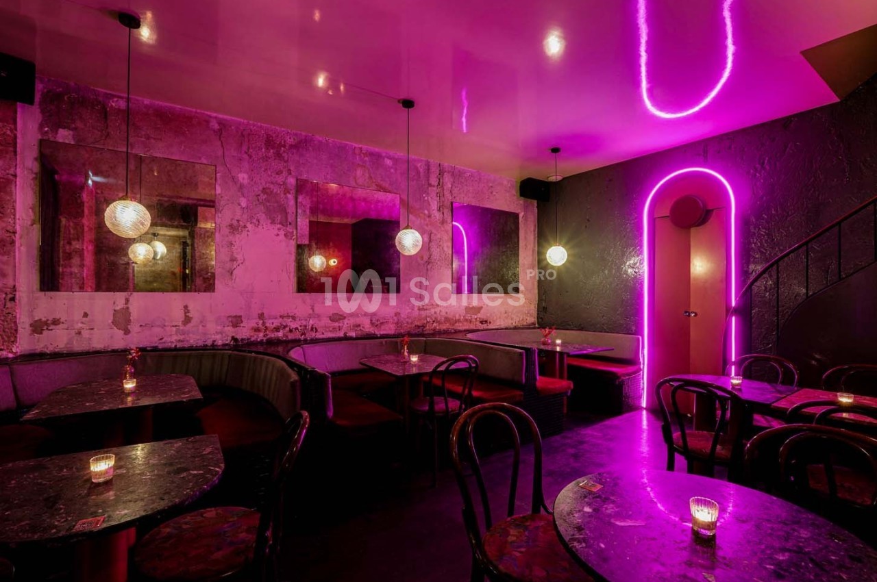 Salle de bar avec éclairage néon rose, murs texturés, banquettes, tables en marbre et chaises en bois.