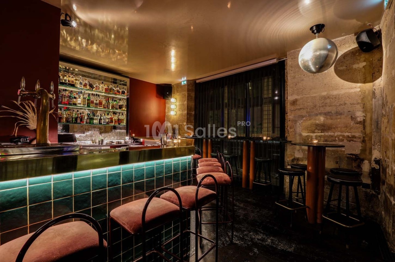 Intérieur d'un bar avec comptoir éclairé, tabourets rembourrés, tables hautes et mur en pierre apparente.