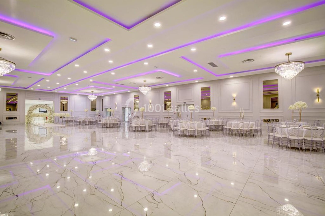 Salle de réception spacieuse avec sol en marbre, tables rondes dressées et éclairage violet au plafond.