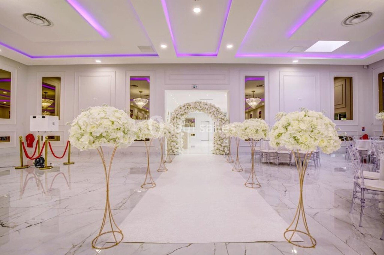 Salle de réception élégante avec allée centrale bordée de bouquets de fleurs blanches et éclairage violet au plafond.