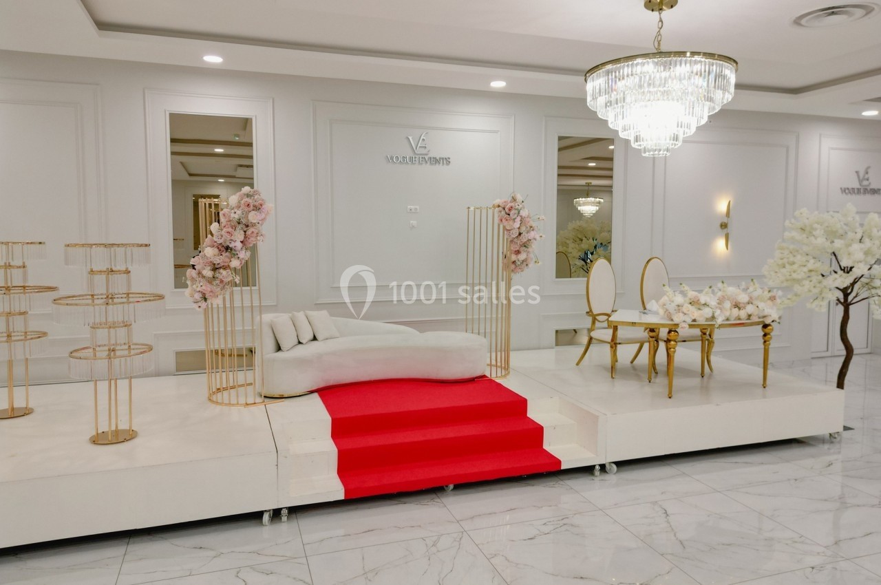 Salle décorée avec un canapé, une table avec chaises dorées, des fleurs et un tapis rouge sur une estrade blanche.