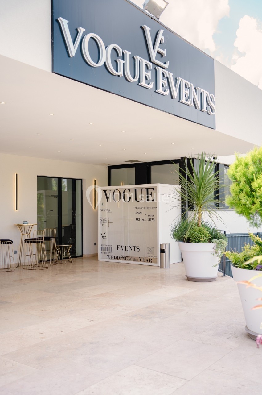 Entrée d'un bâtiment moderne avec un panneau ’Vogue Events’, des plantes et des tables en extérieur.