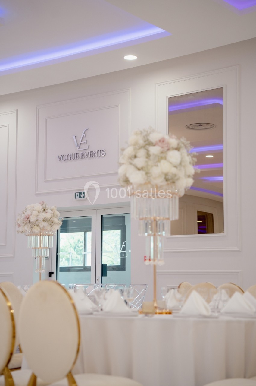 Salle de réception élégante avec tables dressées, chaises dorées et décorations florales blanches sur fond lumineux.