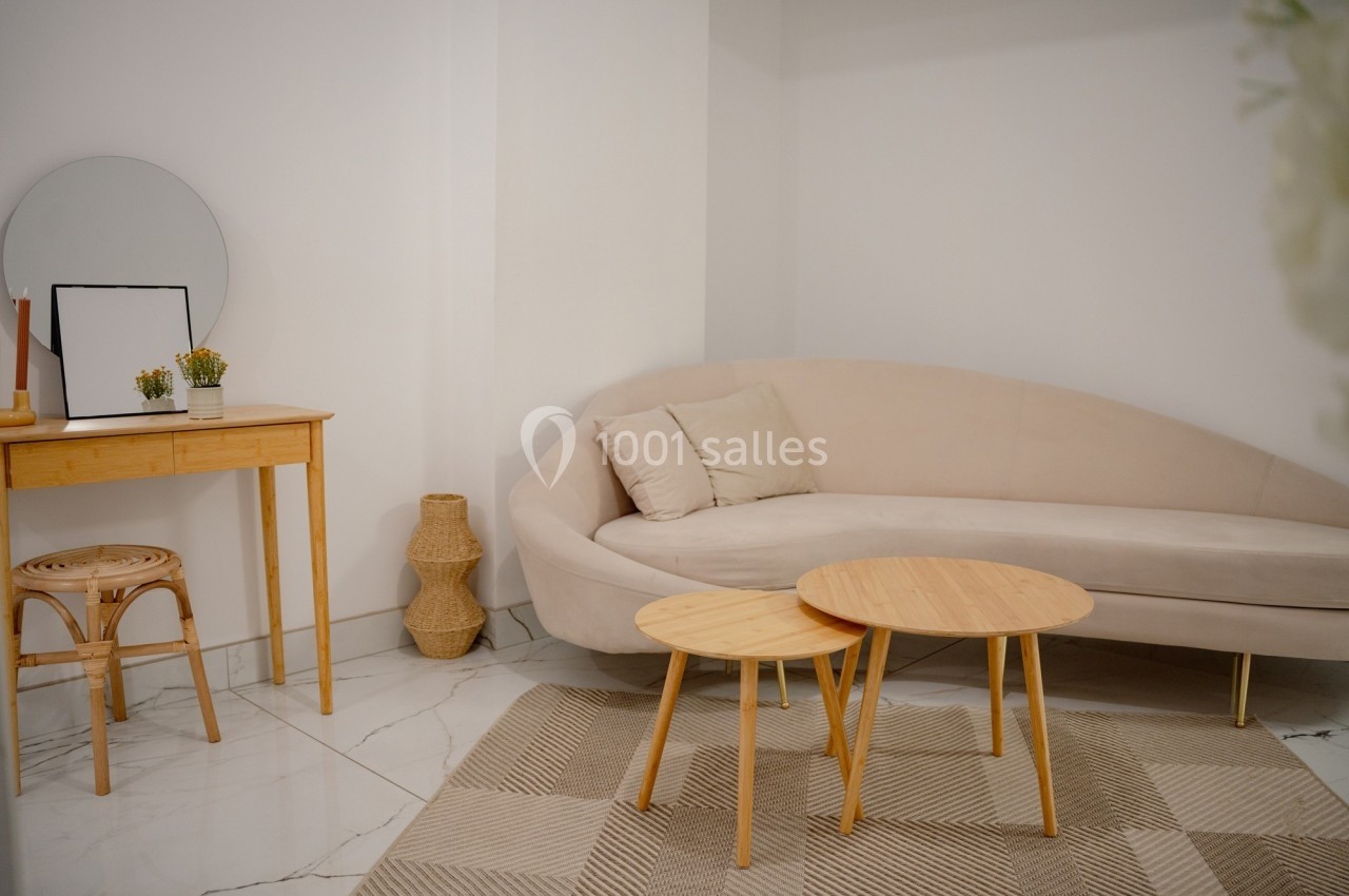 Salon minimaliste avec canapé beige, tables basses en bois clair, console avec miroir et chaise en rotin sur sol en marbre.
