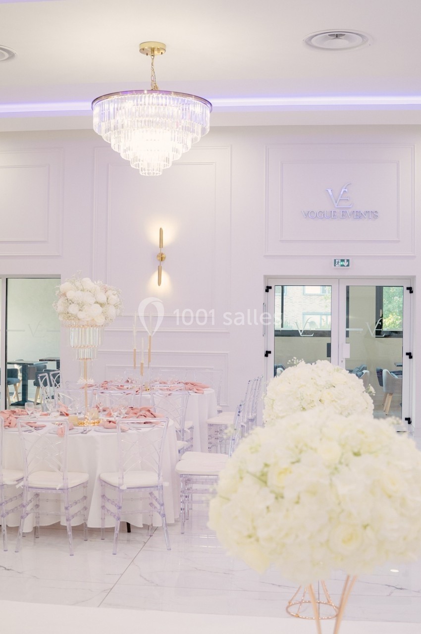 Salle de réception élégante avec tables décorées de fleurs blanches, chaises transparentes et lustre en cristal.