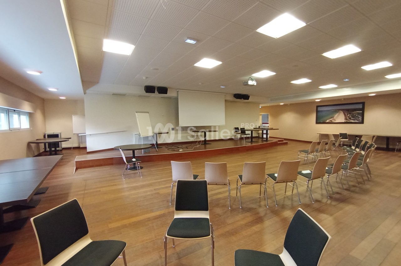 Salle de conférence avec chaises disposées face à un écran de projection, tableau blanc et tables en arrière-plan.