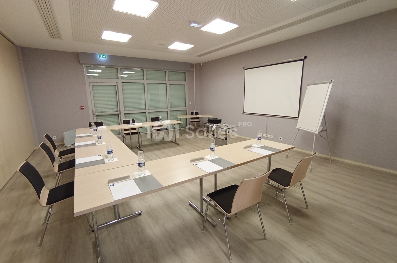Salle de réunion lumineuse avec tables en U, chaises, écran de projection, tableau blanc et fournitures de bureau.