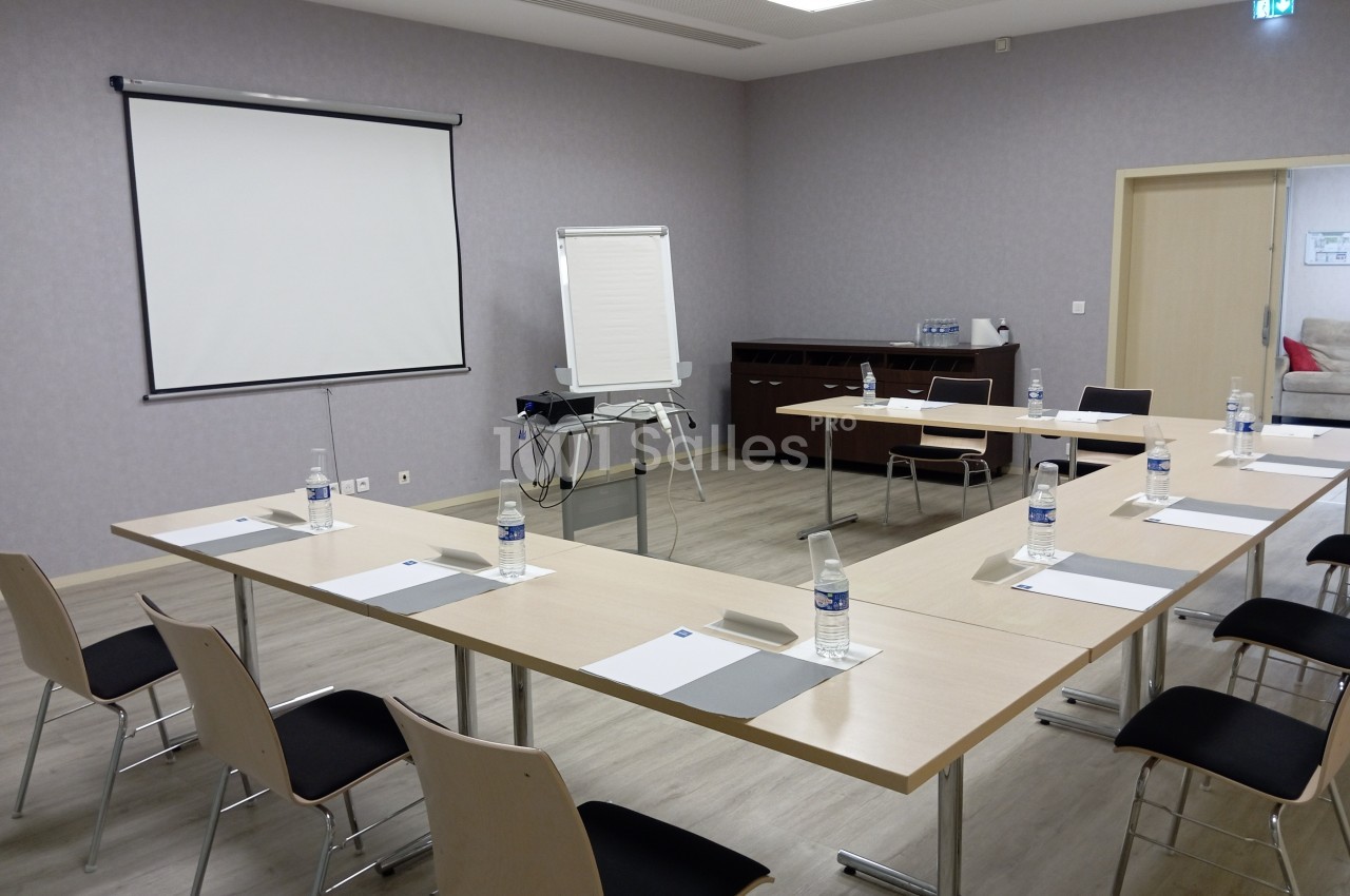 Salle de réunion équipée de tables disposées en U, chaises, paperboard, écran de projection et bouteilles d'eau.