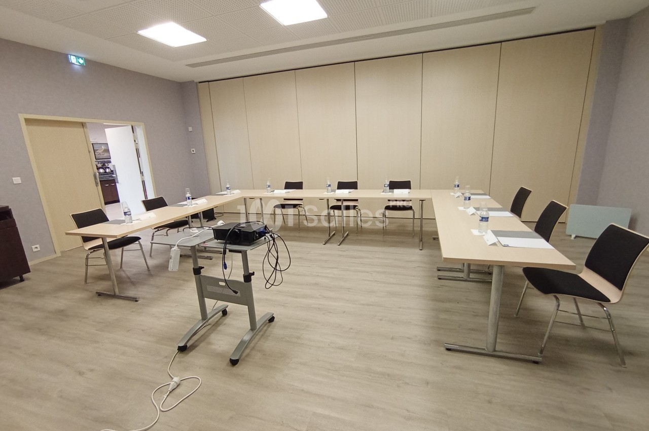 Salle de réunion équipée de tables disposées en U, chaises, bouteilles d'eau et matériel de projection.