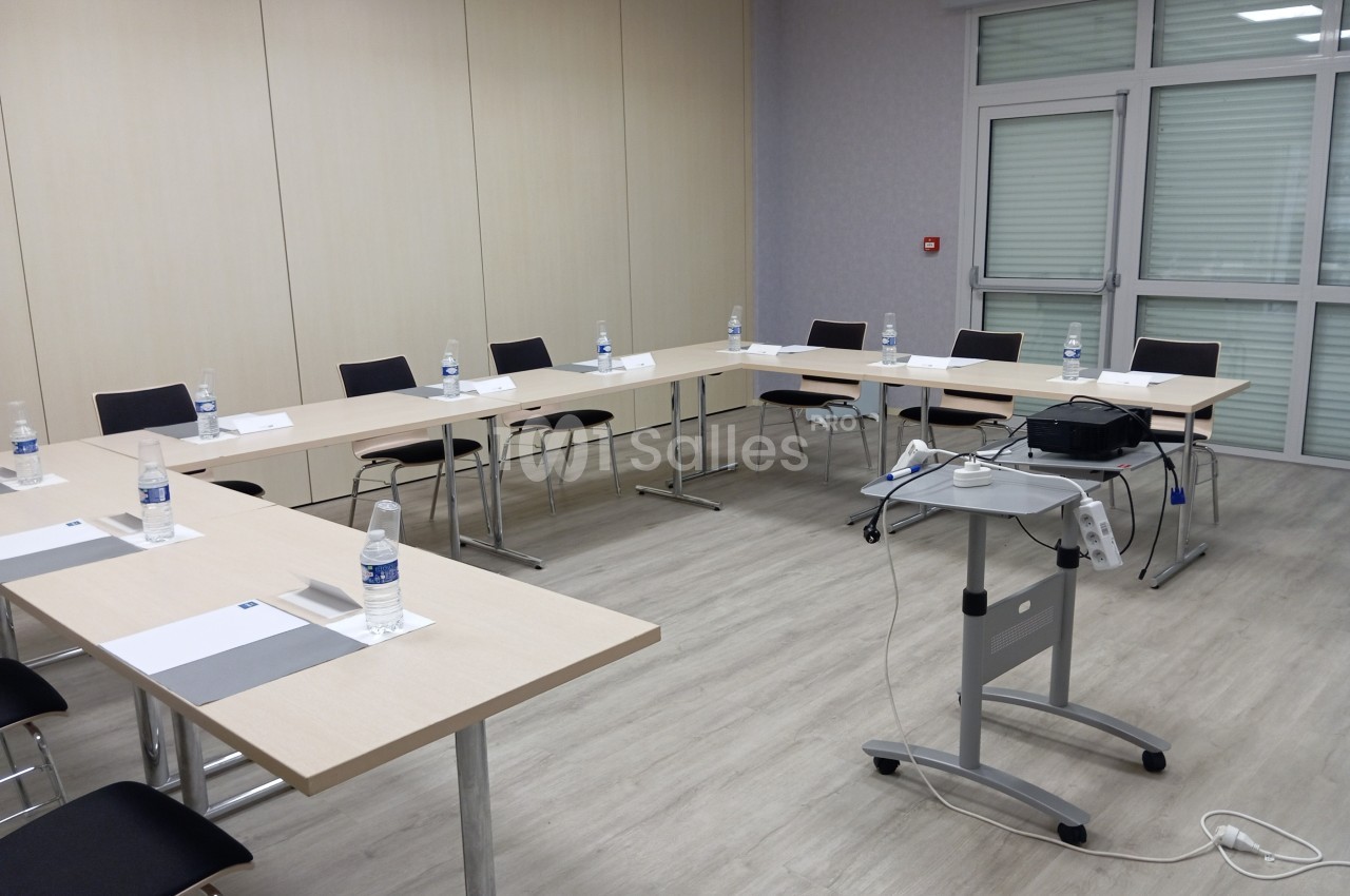 Salle de réunion avec tables disposées en U, chaises, bouteilles d'eau, documents et un projecteur sur support mobile.